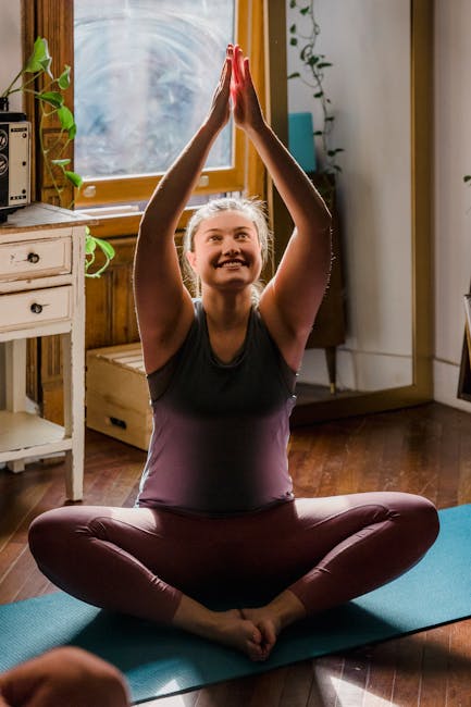 A smiling woman meditating on a yoga mat near a window.
